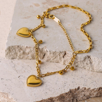 The Ava Grace Necklace is elegantly draped over a beige, textured stone surface. This layered gold necklace features a variety of chains, including a paperclip chain and a beaded chain, all adorned with multiple small and large heart and ball charms.