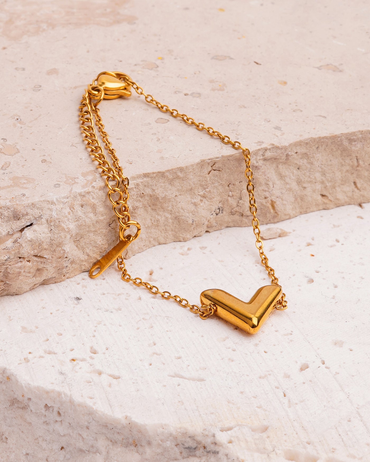 The Avani Arrow Bracelet is showcased on a textured, beige stone surface. The gold-toned bracelet features a delicate chain leading to a prominent, polished V-shaped charm.