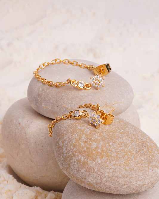 The Azara Chain Studs are showcased on two smooth white stones in a bed of fine white powder. The gold-toned earrings feature a delicate chain that loops from the stud backing to a charm consisting of a small, floral-shaped cluster of gemstones and a single round gemstone.