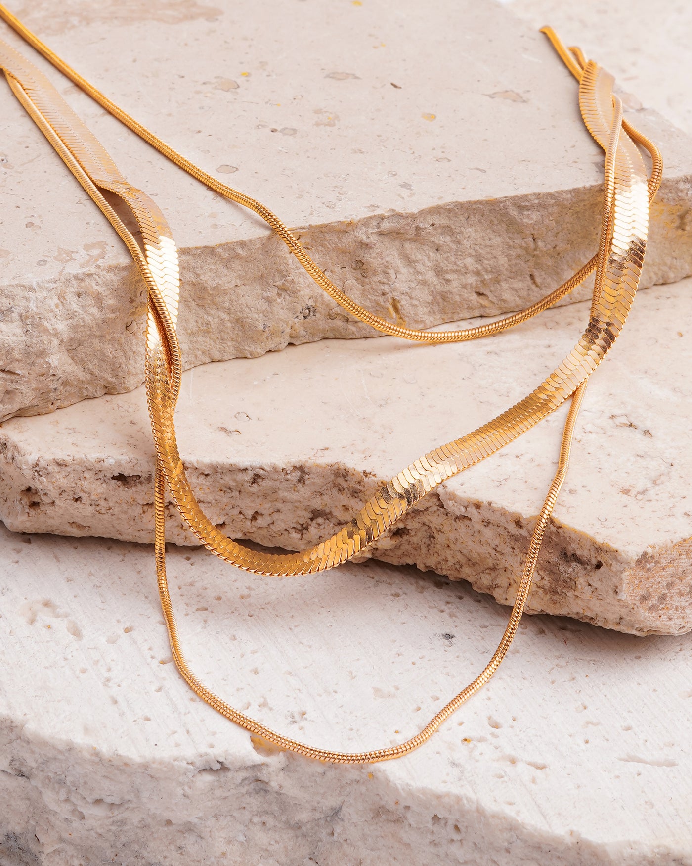 The Cassie Layered Herringbone Necklace is elegantly draped over a textured, beige stone surface. The gold-toned multi-chain necklace showcases its various textures, including a sleek herringbone chain and finer, twisted chains, contrasting beautifully with the rough rock.
