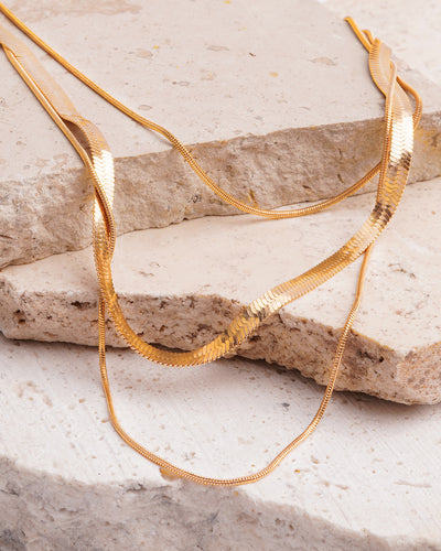 The Cassie Layered Herringbone Necklace is elegantly draped over a textured, beige stone surface. The gold-toned multi-chain necklace showcases its various textures, including a sleek herringbone chain and finer, twisted chains, contrasting beautifully with the rough rock.