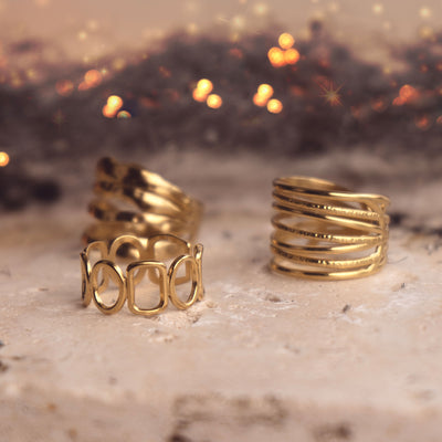 Celeste Radiance Band by Delta Jewel, a modern openwork gold-plated ring, displayed alongside other gold-plated ring designs on a stone surface with a softly glowing background.