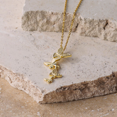 The Celeste Butterfly Necklace is elegantly draped over a beige, textured stone surface. The gold chain leads down to a cascade of four butterfly charms, with some of the butterfly wings shimmering with small, brilliant gemstones.