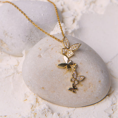 The Celeste Butterfly Necklace rests on a smooth white stone, surrounded by white powder and other stones. The gold-toned necklace features a cascade of four butterfly pendants, with two of them encrusted with small, clear gemstones.