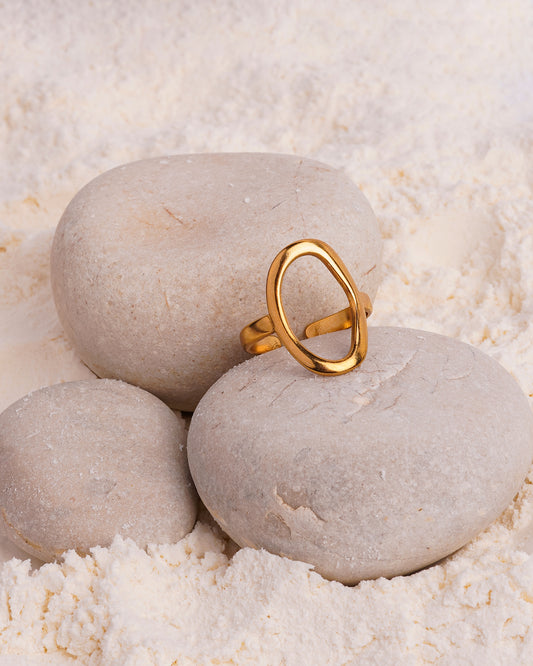 The Chloe Open Teardrop Ring is showcased resting on a smooth white stone in a bed of fine white powder. The gold-toned open ring features a prominent, highly polished oval shape on top.