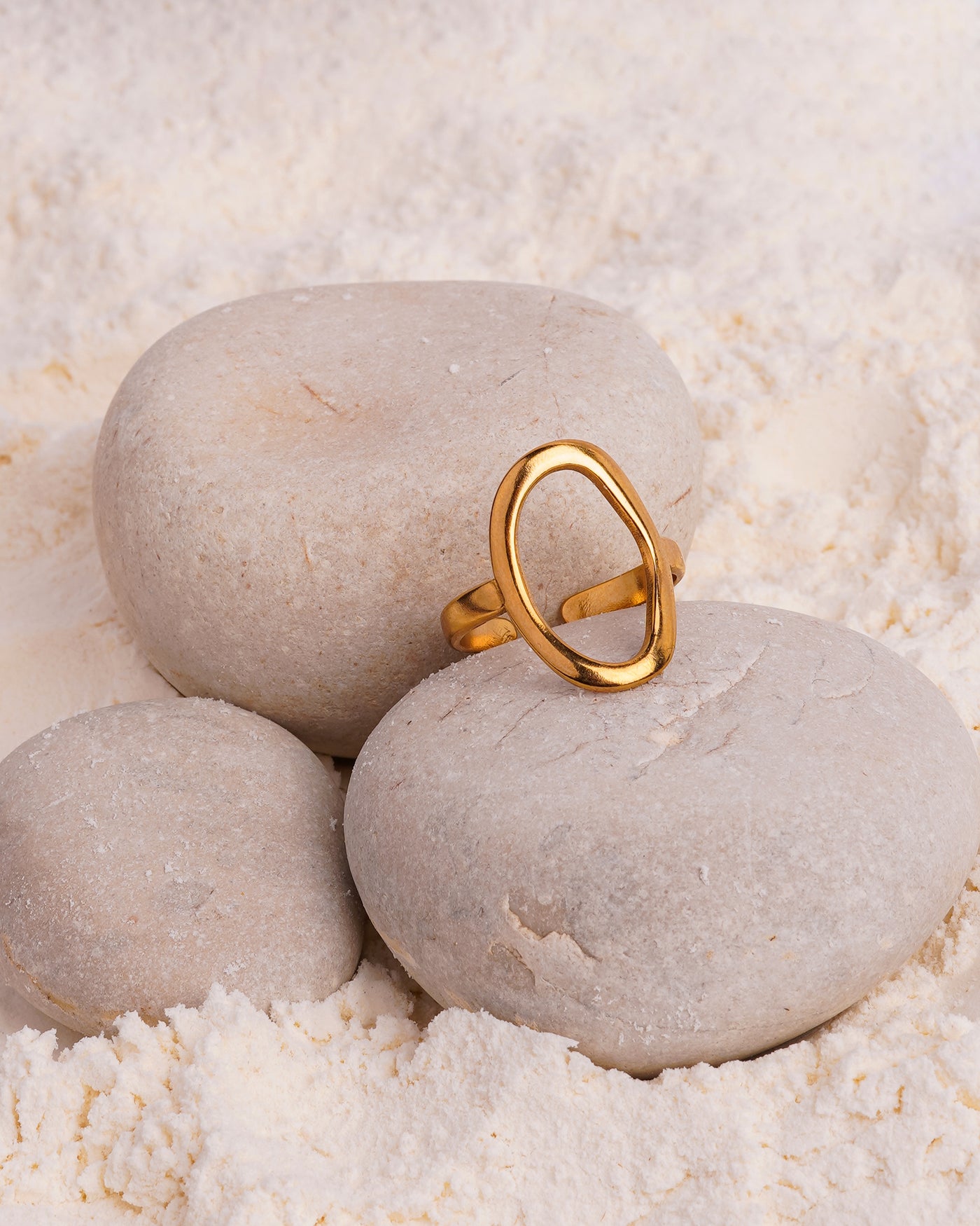 The Chloe Open Teardrop Ring is showcased resting on a smooth white stone in a bed of fine white powder. The gold-toned open ring features a prominent, highly polished oval shape on top.