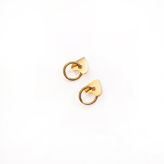 A top-down studio shot against a white background shows a pair of Cora Love Hoops earrings. Each gold-toned earring features a small, polished hoop attached to a solid, flat, and subtly pointed heart-shaped charm.