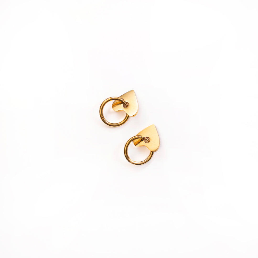 A top-down studio shot against a white background shows a pair of Cora Love Hoops earrings. Each gold-toned earring features a small, polished hoop attached to a solid, flat, and subtly pointed heart-shaped charm.