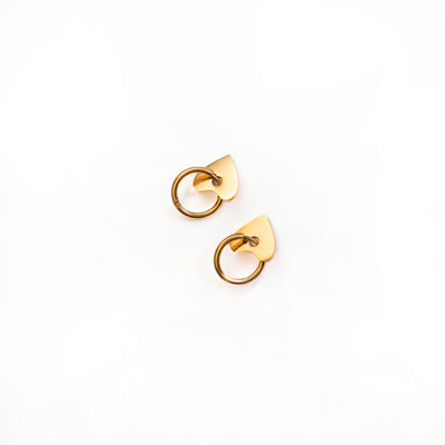 A top-down studio shot against a white background shows a pair of Cora Love Hoops earrings. Each gold-toned earring features a small, polished hoop attached to a solid, flat, and subtly pointed heart-shaped charm.