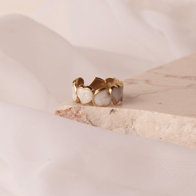  Dahlia Charm Ring by Delta Jewel, featuring a band of pearlescent heart motifs in a gold-plated setting, displayed on a textured stone surface.
