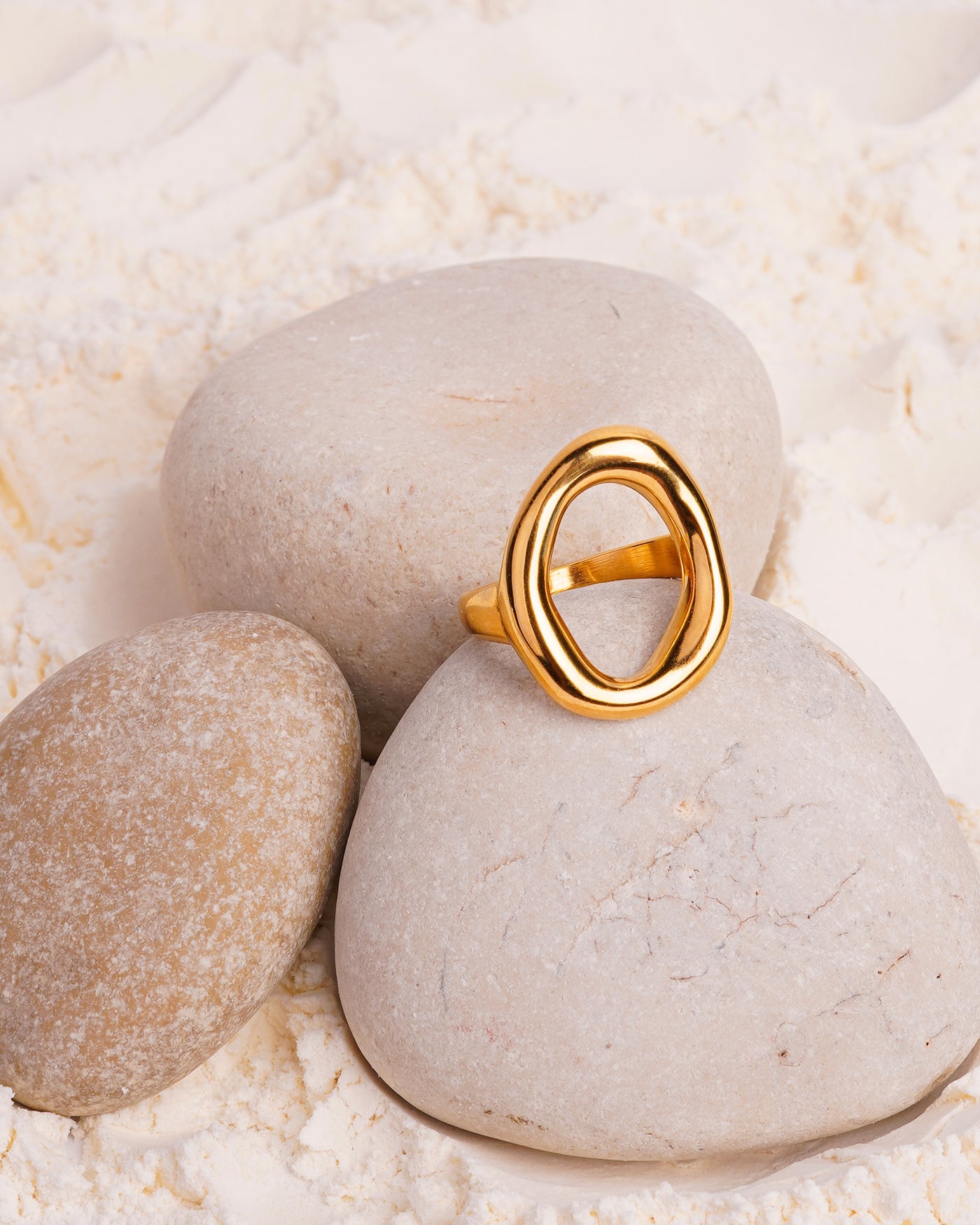 The Elara Infinity Ring is showcased resting on a smooth white stone, surrounded by other stones and a bed of fine white powder. The gold-toned ring has a fluid, intertwining shape that forms an abstract infinity design.