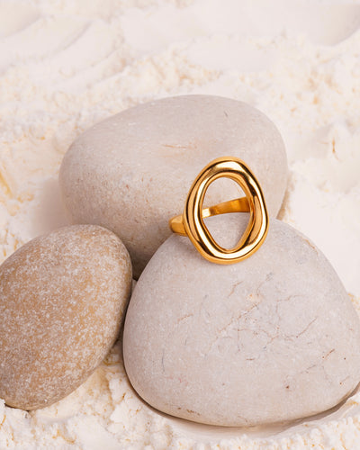 The Elara Infinity Ring is showcased resting on a smooth white stone, surrounded by other stones and a bed of fine white powder. The gold-toned ring has a fluid, intertwining shape that forms an abstract infinity design.