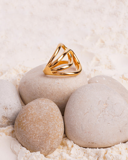 The Elara Wave Ring is showcased resting on a smooth white stone, surrounded by other stones and a bed of fine white powder. The gold-toned ring's fluid, intertwined design is showcased in a still-life shot.