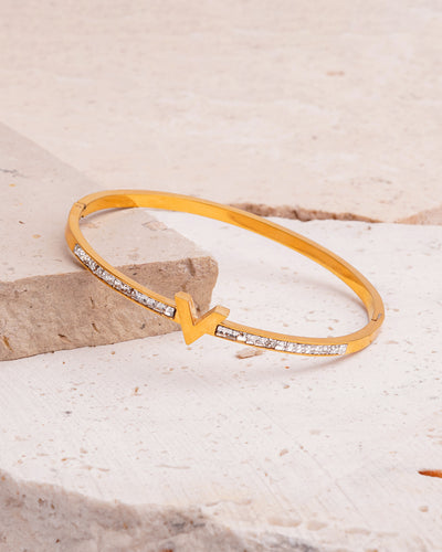 The Elysian Sparkle Bracelet, a gold-toned bangle, is showcased on a beige, textured stone surface. The bangle has a central V-shaped design and is adorned with a line of clear, sparkling gemstones on both sides.