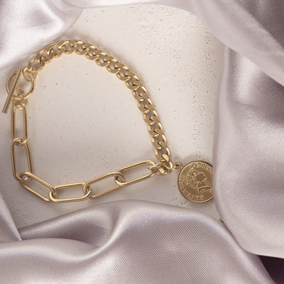 Isolde Luxe Bangle by Delta Jewel, a gold-plated bracelet featuring mixed link styles and a coin charm, displayed on a white surface with satin fabric.