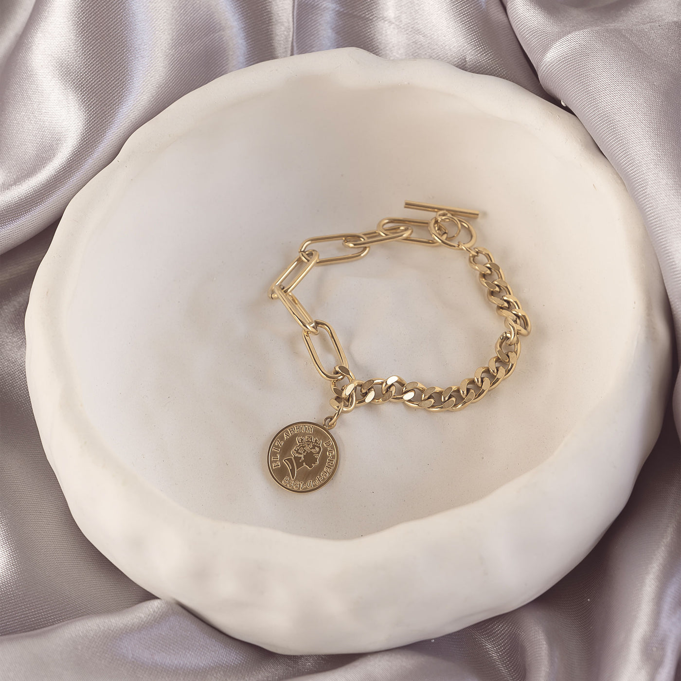 Isolde Luxe Bangle by Delta Jewel, a gold-plated chain bracelet with a toggle clasp and coin detail, elegantly styled in a ceramic dish over satin.