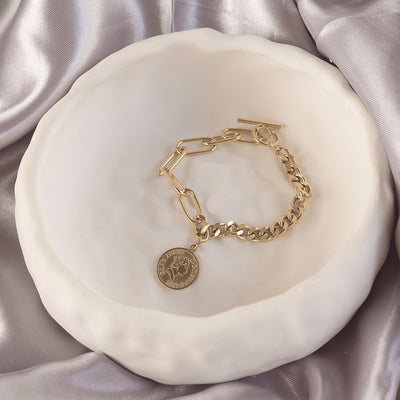 Isolde Luxe Bangle by Delta Jewel, a gold-plated chain bracelet with a toggle clasp and coin detail, elegantly styled in a ceramic dish over satin.