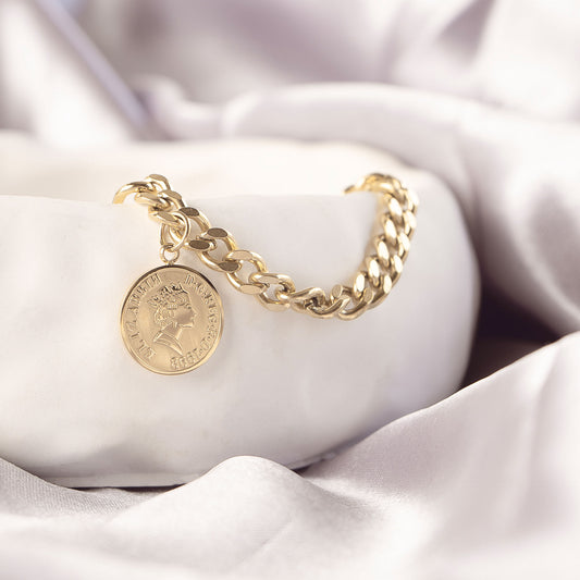 Isolde Luxe Bangle by Delta Jewel, close-up of the gold-plated bracelet highlighting the engraved coin charm, draped over a soft white ceramic dish.