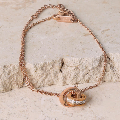 The Ivy Radiance Bracelet is displayed on a beige, textured stone surface. This rose gold-toned bracelet has a delicate chain connected to a centerpiece of two interlocking rings, one with Roman numerals and the other with sparkling cubic zirconia.