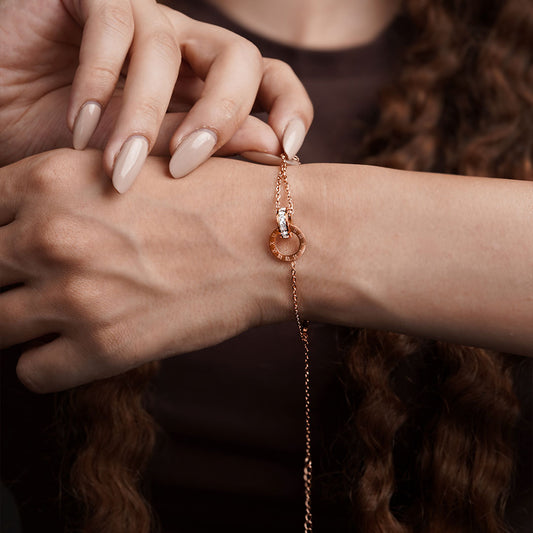 A close-up of a person's wrist and hand, showing the Ivy Radiance Bracelet. The rose gold-toned chain leads to a centerpiece of two interlocking rings; one is engraved with Roman numerals, and the other is covered in small, clear gemstones.