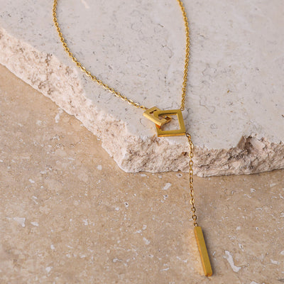 Gold Plated Jasmine Grace Necklace with two interlocking squares, with one forming a lariat and bar drop pendant. The necklace is displayed on a textured beige and cream-colored stone surface.
