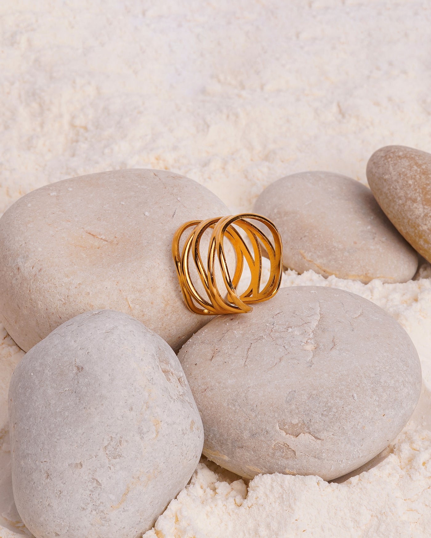 The Lia Lace Ring rests on a large, smooth white stone, surrounded by other stones and a bed of fine white powder. The gold-toned open ring's bold, intertwined band design is showcased in this still-life shot.