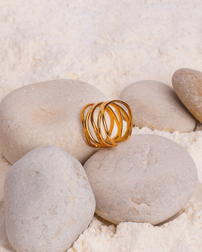 The Lia Lace Ring rests on a large, smooth white stone, surrounded by other stones and a bed of fine white powder. The gold-toned open ring's bold, intertwined band design is showcased in this still-life shot.