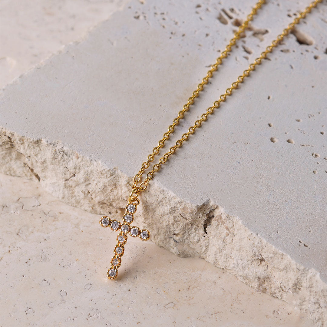 The Luminous Faith Pendant, a gold-toned necklace, is showcased on a rugged, light-colored stone surface. The cross pendant is the focal point, encrusted with a line of clear, sparkling gemstones.