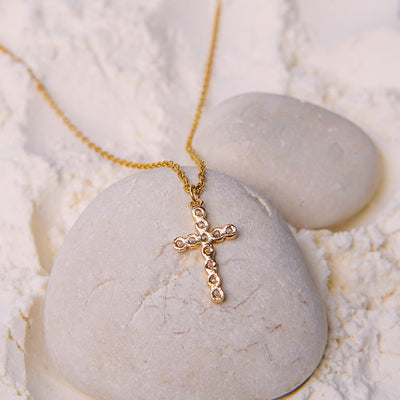 The Luminous Faith Pendant rests on a smooth white stone, surrounded by fine white powder. The gold chain of the necklace leads to the cross pendant, which is adorned with numerous tiny, glittering clear gemstones.