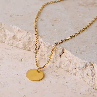 The Luna Disc Necklace rests on a rugged, light-colored stone surface. Its delicate gold-toned chain contrasts with the textured rock, leading to a simple, polished circular pendant that reflects the light.