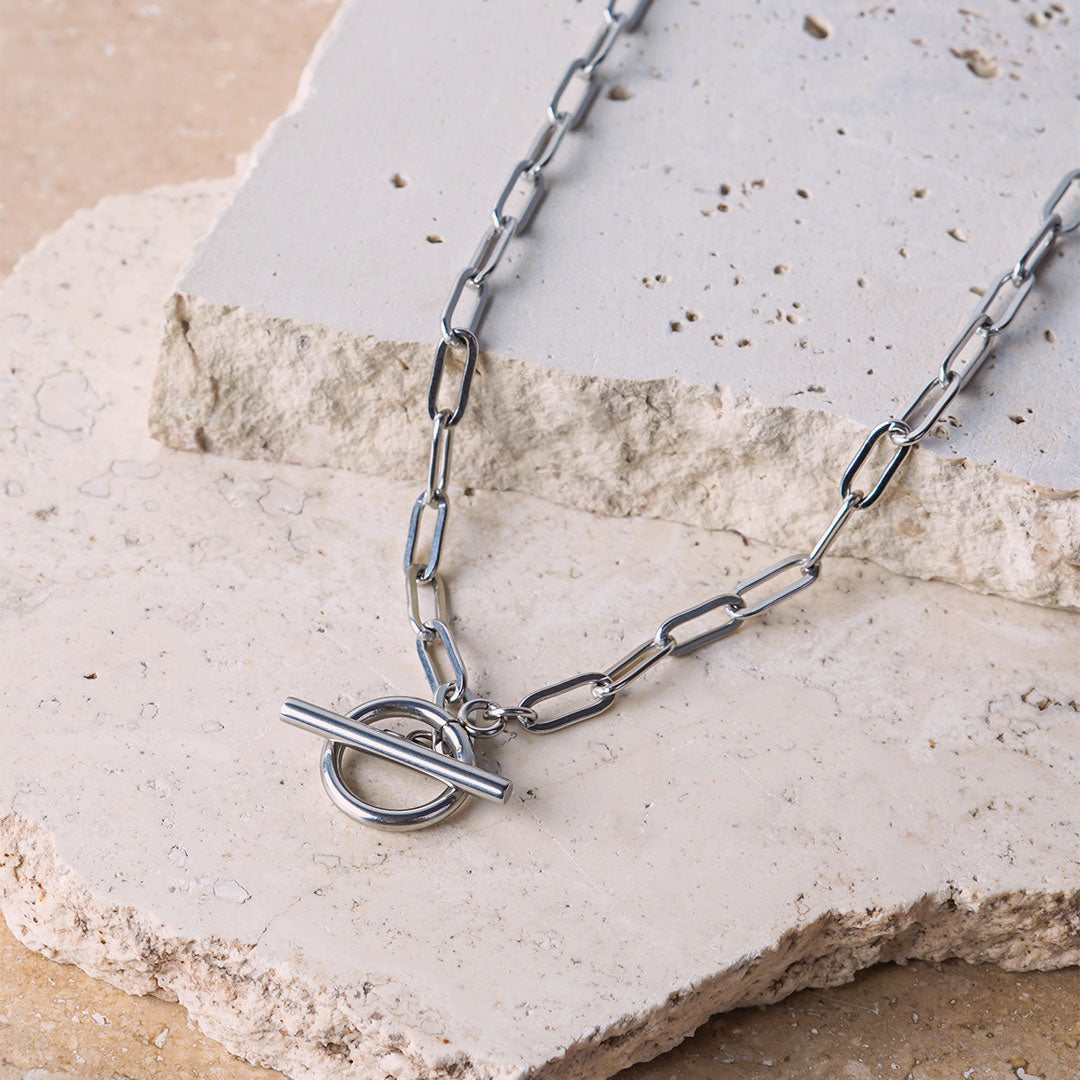 The Lustrous Arc Necklace is displayed on a light-colored, textured stone surface. This silver-toned necklace features a chain of elongated links, with a large, polished toggle clasp at its center.