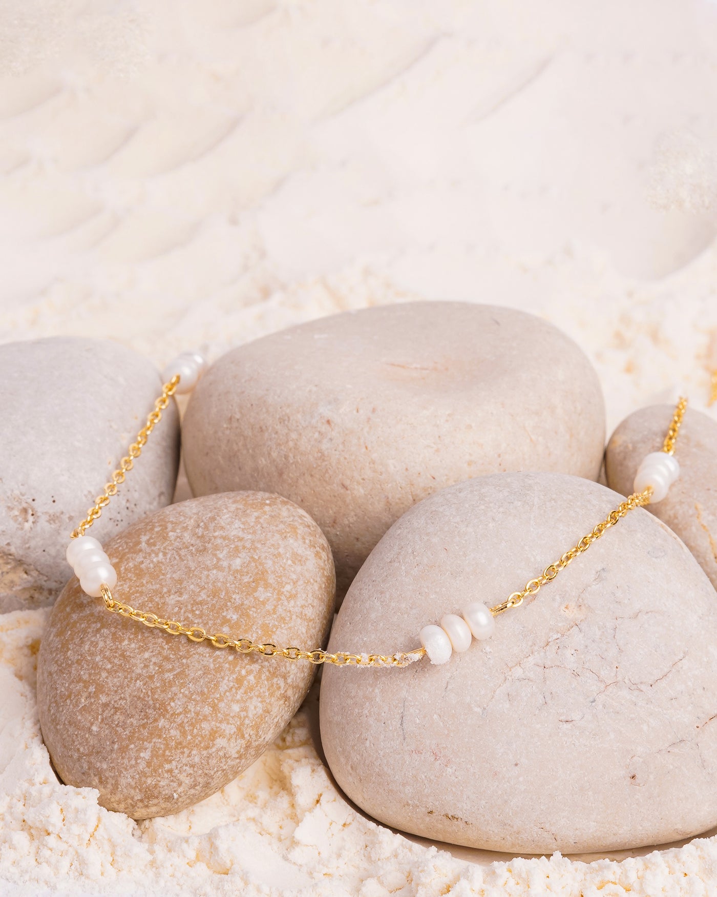The Lyra Pearl Chain Necklace is elegantly arranged on a bed of fine white powder, resting on a cluster of smooth, light-colored stones. The gold-toned chain is adorned with several small groups of white pearls.