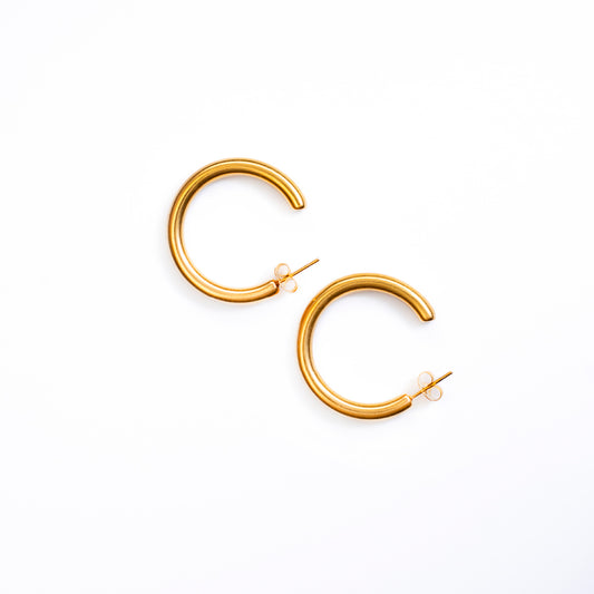 A top-down studio shot on a clean white background displays a pair of Marina Bold Hoops earrings. Each earring is a substantial, highly polished, and perfectly curved gold-toned hoop with a post backing.