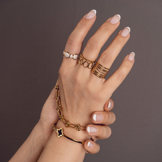 Ophelia Gleam Cuff by Delta Jewel, a slim gold-plated bracelet featuring a black clover charm, worn with layered gold jewelry on the wrist and fingers.