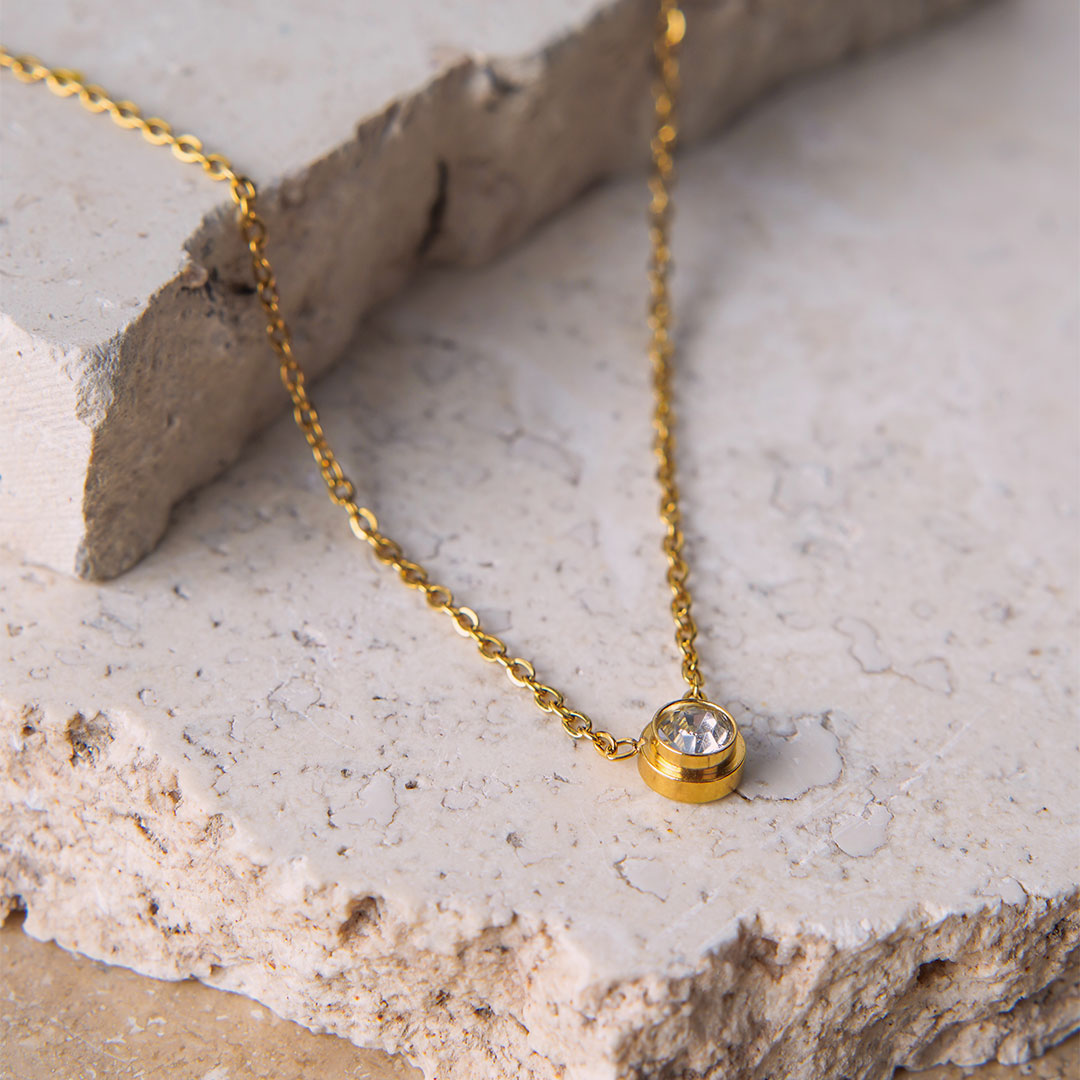 The Parker Spheroid Necklace is showcased on a beige, textured stone surface. The gold-toned necklace features a fine chain that gracefully suspends a single, round, bezel-set cubic zirconia pendant, catching the light beautifully.