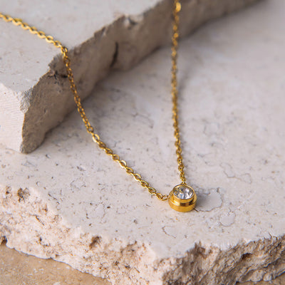 The Parker Spheroid Necklace is showcased on a beige, textured stone surface. The gold-toned necklace features a fine chain that gracefully suspends a single, round, bezel-set cubic zirconia pendant, catching the light beautifully.