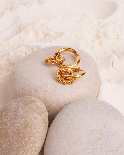 The Penelope Knot Hoops are showcased resting on two smooth white stones in a bed of fine white powder. The gold-toned earrings feature a polished hoop with a smaller, intricately knotted ring dangling from it.