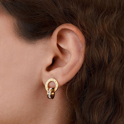 A close-up side profile shows a person's ear wearing a Roma Timeless Hoop earring. The gold-toned earring, featuring two interlocking rings (one with Roman numerals, the other with gemstones), hangs elegantly from the earlobe.