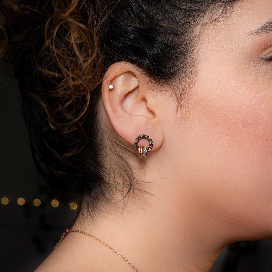 A close-up side profile shows a person's ear wearing a Roma Timeless Hoop earring. The gold-toned earring, featuring two interlocking rings (one with Roman numerals, the other with gemstones), hangs elegantly from the earlobe.
