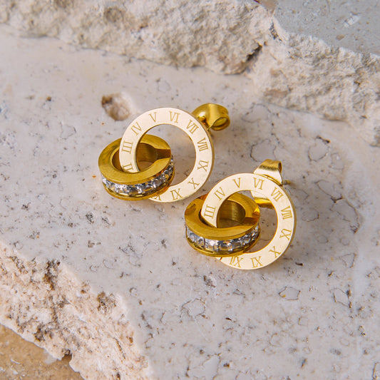 A pair of Roma Timeless Hoops are displayed on a beige, textured stone surface. Each gold-toned earring consists of two interlocking rings: one with Roman numeral engravings and the other encrusted with clear gemstones.