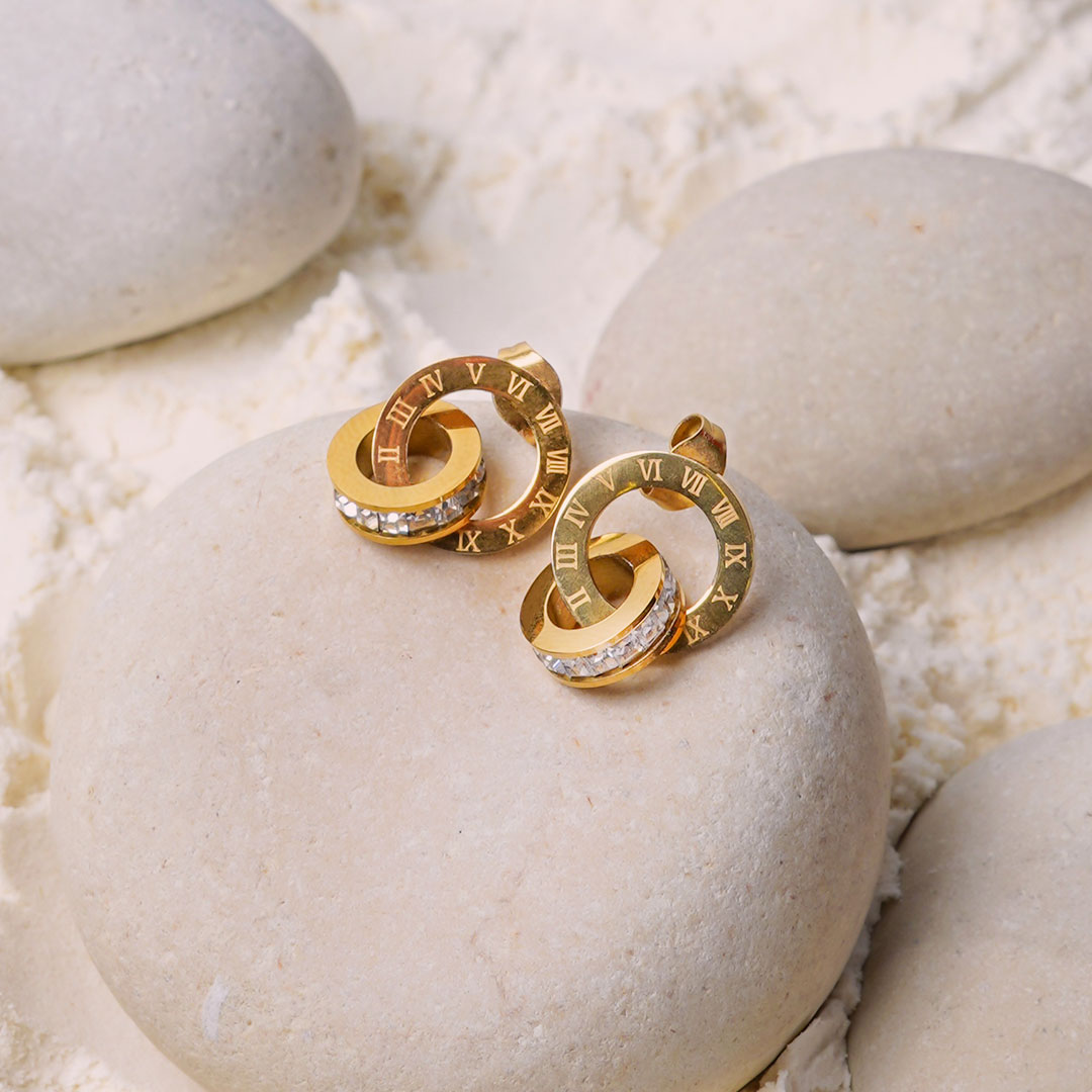 The Roma Timeless Hoops rest on a smooth white stone, surrounded by white powder. This pair of gold-toned earrings showcases a design of two interconnected rings, one with Roman numerals and the other with a line of sparkling gemstones.