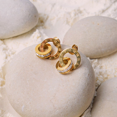 The Roma Timeless Hoops rest on a smooth white stone, surrounded by white powder. This pair of gold-toned earrings showcases a design of two interconnected rings, one with Roman numerals and the other with a line of sparkling gemstones.