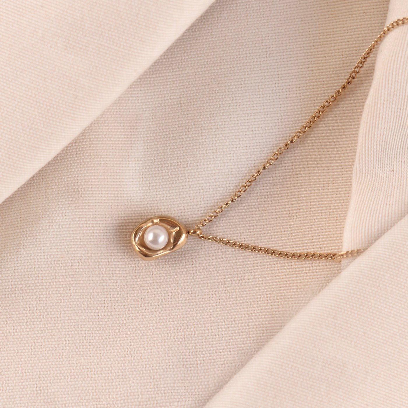 The Saffron Luxe Necklace by Delta Jewel, made from high-quality stainless steel with a luxurious gold plating, featuring an abstract pendant cradling a pearl, displayed elegantly on soft beige fabric.