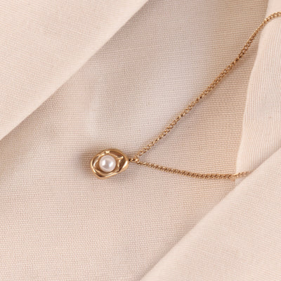 The Saffron Luxe Necklace by Delta Jewel, made from high-quality stainless steel with a luxurious gold plating, featuring an abstract pendant cradling a pearl, displayed elegantly on soft beige fabric.