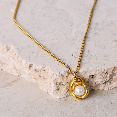 The Saffron Luxe Necklace is showcased on a beige, textured stone surface. The gold-toned necklace features a curb chain that leads to a unique, organically shaped pendant with a single white pearl set at its center.