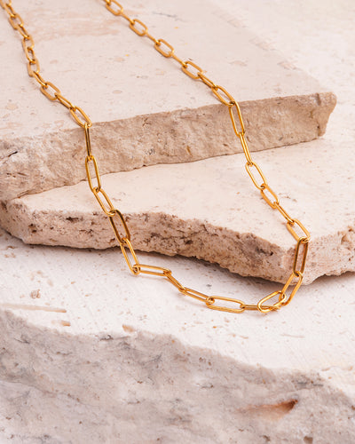 The Serena Paperclip Necklace is showcased on a beige, textured stone surface. The gold-toned necklace features a chain of sleek, elongated links that are draped across the rock.