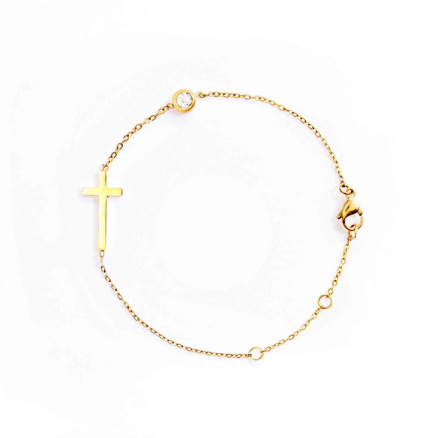 The Stella Grace Bracelet is presented in a studio shot on a white background, forming a circle. This gold-toned bracelet features a minimalist, flat cross charm positioned along the chain, with a small, clear round gemstone set further along the chain.