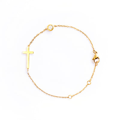 The Stella Grace Bracelet is presented in a studio shot on a white background, forming a circle. This gold-toned bracelet features a minimalist, flat cross charm positioned along the chain, with a small, clear round gemstone set further along the chain.