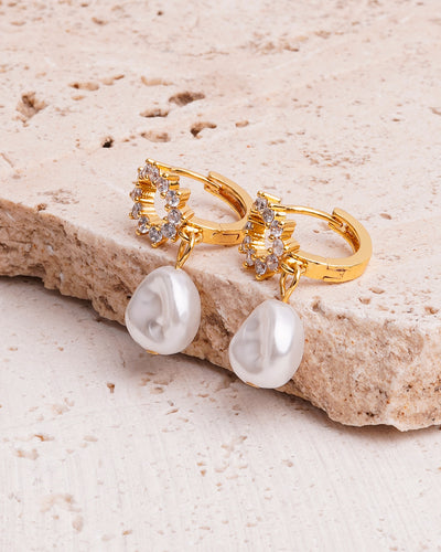 A pair of Stella Regal Drops earrings are showcased on a beige, textured stone surface. The gold-toned earrings feature a top charm encrusted with clear gemstones and a large, irregularly shaped white pearl hanging below.