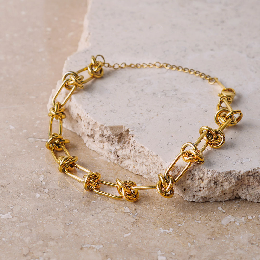  The Valerie Knotted Bracelet is showcased on a beige, textured stone surface. The gold-toned bracelet features a series of elongated links connected by intricate, polished knots, creating a bold and sculptural design.